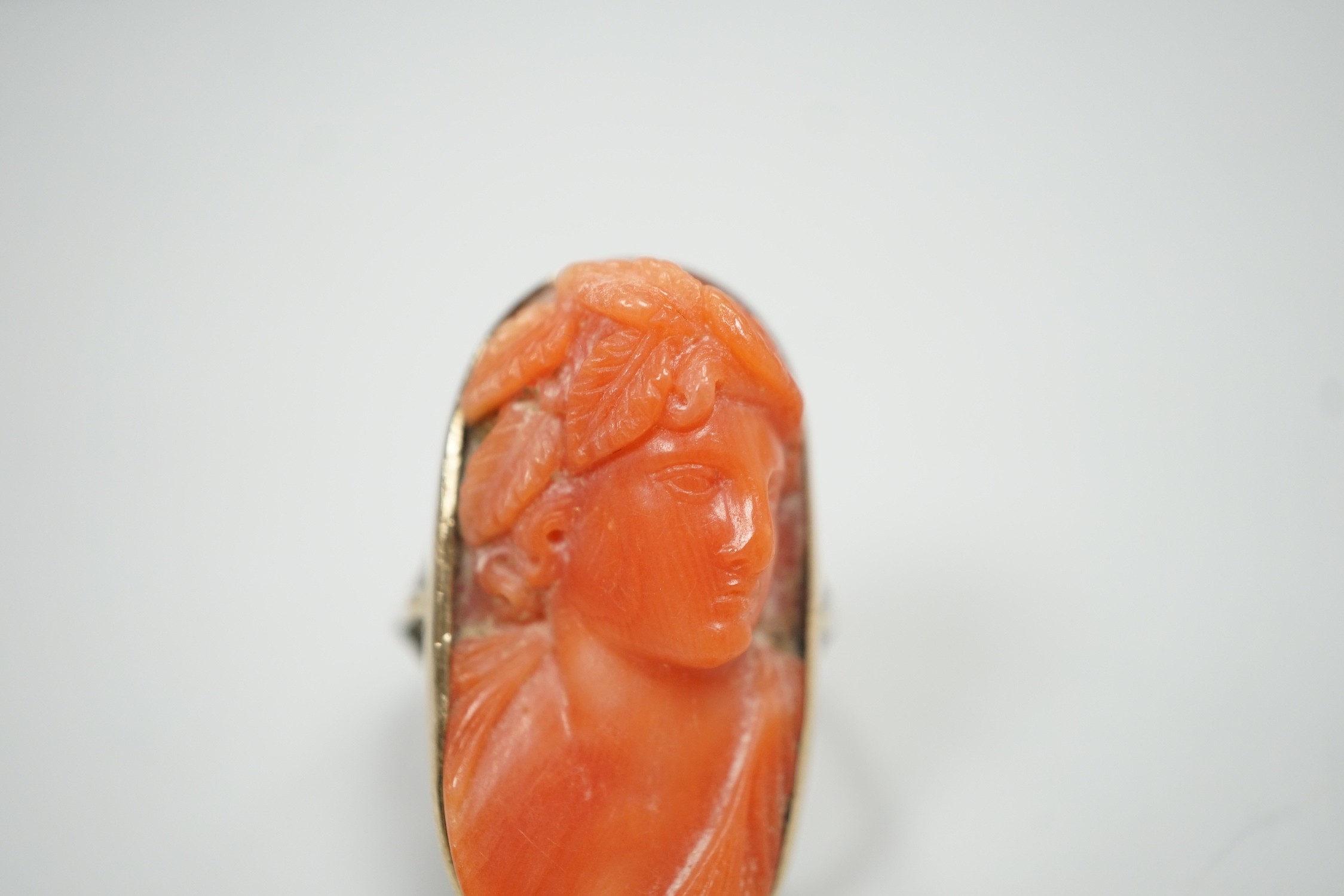 A yellow metal and coral set oval dress upfinger ring, the coral carved with a classical bust to sinister, size O, gross weight 8.2 grams.